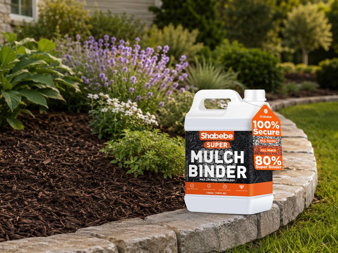 A bottle of Shabebe Super Mulch Binder stands on a stone border in a landscaped garden bed with mulch, greenery, and purple flowers in the background.